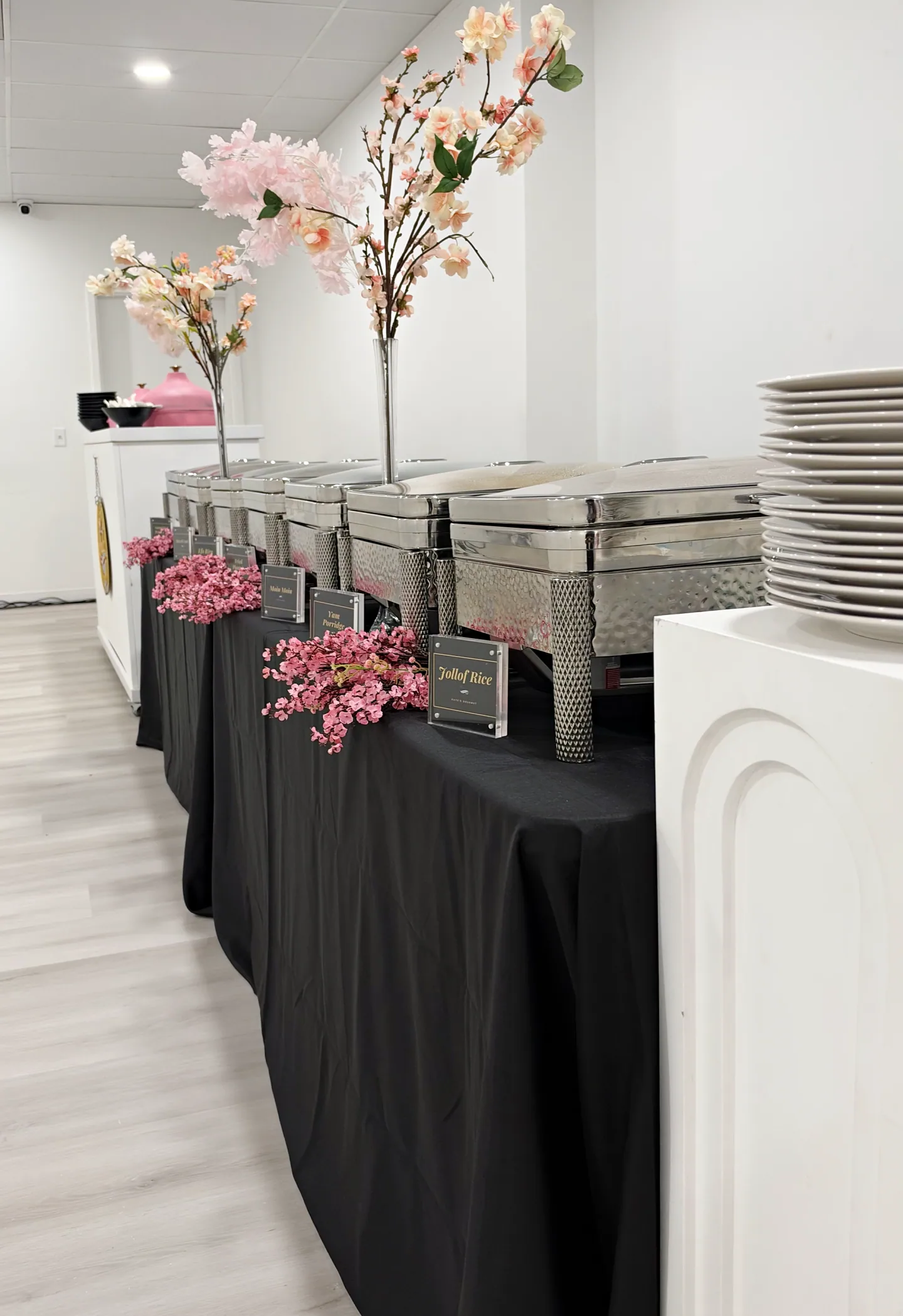 A buffet station with silver chafing dishes, pink flowers, and stacked plates.
