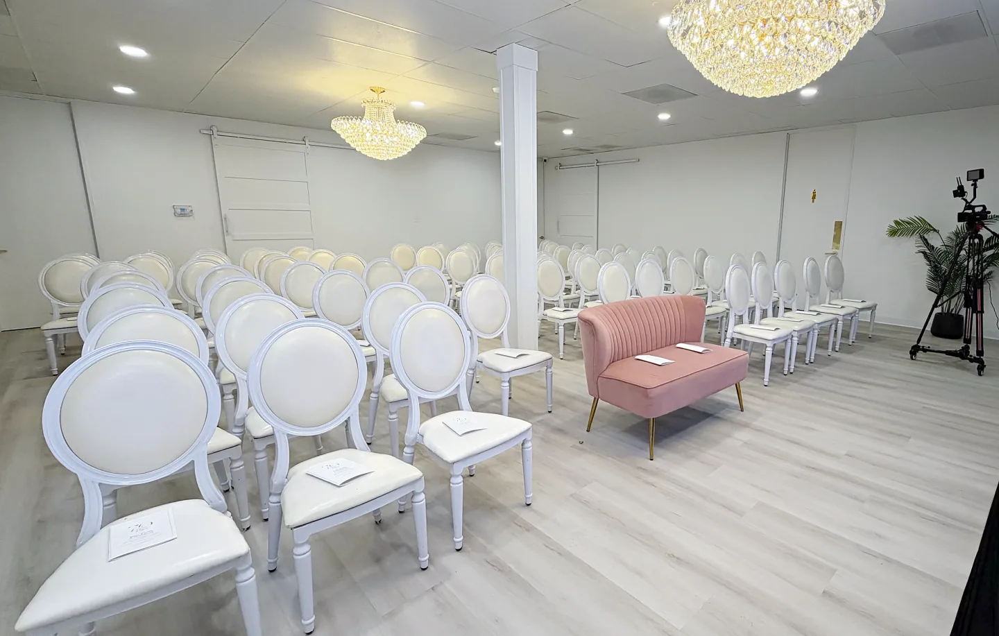 Rows of white chairs and a blush loveseat beneath chandeliers in the cocktail area.