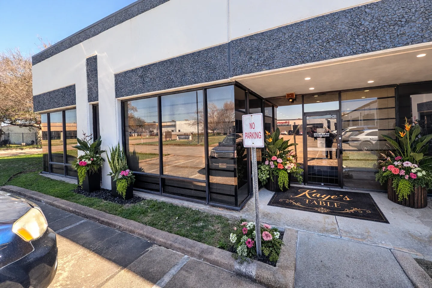 The daytime entrance to Kaye’s Table with flower-filled planters and a branded entry mat.